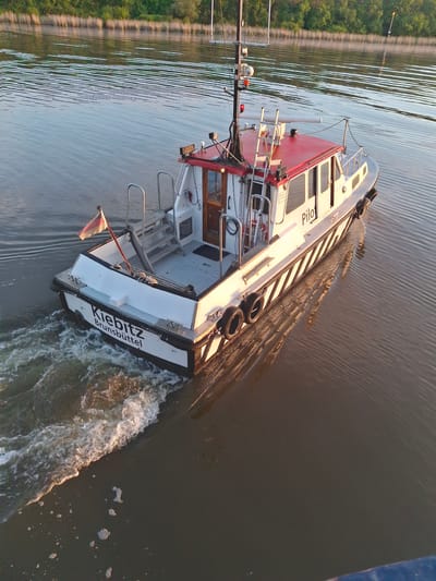 A color, high-angle, outdoor photograph depicts a white and red pilot boat named “Pilot Kiebitz Brunsbüttel” traveling through a body of water, possibly a river or canal, in Brunsbüttel, Germany.

The boat, which is tilted toward the right, is white with a red roof. The word "Pilot" is seen on the side of the boat, while "Kiebitz Brunsbüttel" is written on the back. Tires are hung on the side of the boat. A flag is visible at the stern.

The boat is moving, as evidenced by the white water being churned up behind it. The water appears calm with slight ripples. The boat is surrounded by a body of water. A shoreline with green trees is seen in the background. The light suggests it might be early morning or late afternoon.

A blue object is seen in the upper right corner of the photo, but is out of focus.