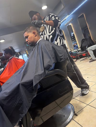 The image captures an active scene within a modern barbershop or hair salon, located in Blouberg, South Africa. The photograph is taken from a high, tilted perspective, showcasing two clients and a barber.
In the center, a male client is seated in a black professional barber chair, draped in a dark grey barber cape. He has a fade haircut and a beard, appearing calm during his grooming session. To his left, the barber is standing, holding a small brush, likely in the process of tidying up loose hair after a cut. The barber wears a black cap, a black and white patterned long-sleeve shirt, a watch on his left wrist, and black gloves.
On the tiled floor around the barber chair, hair clippings are visible, along with a silver basin collecting more hair, indicating active grooming. In the lower right foreground, a second client, wearing an orange cape and sporting dreadlocks, is partially visible, suggesting multiple services are underway.
The barbershop features a light-colored tiled floor and a modern ceiling equipped with recessed circular lights and linear LED strip lighting. Reflective panels on the ceiling offer glimpses of other areas of the shop and additional individuals. Prominently displayed on one of the ceiling beams, illuminated by a strip light, is the white numeric text "0612169721," likely a contact number for the establishment. The client in the chair is wearing distressed black/grey jeans and grey and white sneakers. The overall setting appears clean, well-lit, and professional.