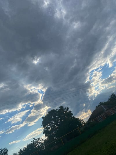 The image shows a dramatic sky filled with dark, heavy clouds interspersed with patches of bright blue sky illuminated by sunlight. Rays of light pierce through the clouds in some areas. The perspective is from ground level, looking upwards and slightly to the right. In the foreground, there is a grassy slope with a yellow-topped fence. To the right, a small, rustic house with a corrugated roof is visible, partially obscured by trees. A large, dark tree is prominent in the middle ground. Power lines stretch horizontally across the upper portion of the image. The scene appears to be in an outdoor, possibly rural or suburban, setting. There are no people visible in the image. The environment suggests a possibly overcast day with the sun breaking through.