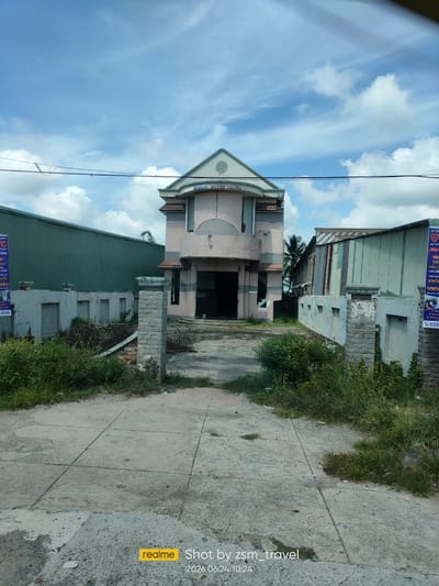 The image shows a large, two-story building with a pink facade, located in the city of Gò Dầu, Vietnam. The building appears to be abandoned or under construction, with boarded-up windows and an open entrance. In front of the building, there is a paved driveway leading to the entrance, flanked by overgrown vegetation and a low concrete wall. To the left and right of the building are large, industrial-style structures, one with a green metal exterior and the other with a corrugated metal exterior. The sky is blue with scattered white clouds. The foreground of the image shows a concrete pathway. A watermark at the bottom right of the image reads "realme Shot by zsm_travel 2026.06.24 10:24".