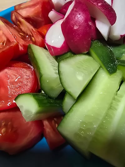 A close-up, overhead shot shows a vibrant blue bowl filled with a salad of chopped tomatoes, cucumbers, and radishes. The tomatoes are cut into wedges, revealing their juicy red interiors. Slices of cucumber, some with visible seeds, are scattered throughout, their light green flesh glistening. Bright pink and white radishes, also sliced, add a contrasting color and texture to the mix. The salad appears fresh, with some water droplets visible on the vegetables, suggesting it may have been recently washed or dressed. The background is blurred, but the edge of a blue bowl is visible, and a hint of a table surface. The scene is brightly lit, likely from overhead natural or artificial light, highlighting the textures and colors of the fresh produce. There is no indication of the location of Ермолаевский Затон, Russia in this image.