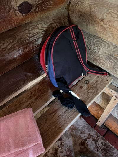 A black and red duffel bag rests on a wooden bench, with a pair of black socks lying beside it. To the left of the bag, a folded pink towel is visible. The bench itself is made of rough, unfinished wood and is situated against a wall constructed from large, unvarnished logs, some of which show prominent knots. The floor beneath the bench is covered with a patterned rug and appears to be made of wood planks, with some red brick or tile visible in the lower right corner. The scene is dimly lit, suggesting an indoor environment. The location is specified as Pobedinka, Russia.