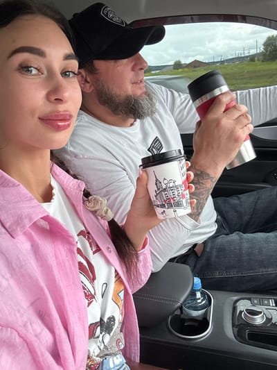 A woman and a man are in a car. The woman is in the foreground, looking at the camera, while the man is in the background, looking forward. The woman is wearing a pink shirt and a white t-shirt. She has long dark hair and is holding a coffee cup. The man is wearing a black baseball cap and a white t-shirt. He has a beard and is holding a red thermos. The car interior is visible, including the dashboard and a cup holder with a bottle of water. The background outside the car shows a grassy landscape with some industrial structures and a cloudy sky. The image appears to have been taken in Pervomaiskoe, Russia.