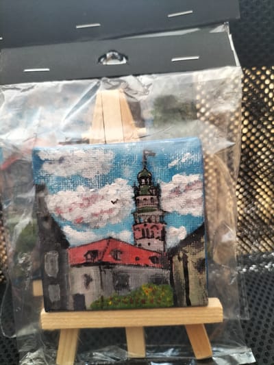 A miniature acrylic painting on canvas depicts the Maidens' Tower (Dívčí věž) in České Budějovice, Czechia. The tower, with its distinctive green roof and flagpole, is rendered in detail, surrounded by stylized buildings with red roofs and grey facades. A patch of green foliage with red and yellow flowers is painted at the base of the tower. The sky is a vibrant blue with fluffy white and pink clouds. The painting is presented on a small wooden easel, encased in clear plastic. The background shows a textured dark surface.