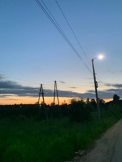 The image depicts a rural landscape at dusk in Ganino, Russia. The scene is dominated by a clear blue sky that transitions into a warm orange and pink sunset near the horizon, with scattered grey clouds. Three power lines stretch diagonally across the left side of the frame, converging towards the top.

In the center, a tall utility pole with an illuminated lamp at its top stands prominently. Further back, two sets of wooden power line poles, forming an 'A' shape, are visible against the sunset. These poles support additional power lines, which are less distinct than those in the foreground.

The right side of the image shows a grassy field bordering a dirt road. Beyond the field, a dense line of dark green trees creates a silhouette against the colorful sky. The overall impression is one of tranquility and the peaceful transition from day to night in a natural setting. The lighting suggests it is either sunrise or sunset, with the lamp already on, indicating twilight. No people or text are visible in the image.