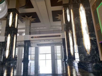 The image shows the interior of a large, modern building, likely a mosque, in Aluekambuk, Indonesia. The space features grand marble pillars with integrated lighting that casts a warm glow. The floor is highly polished, reflecting the light from the pillars and the expansive windows, creating a shimmering effect. A single person is visible in the distance, standing near a window. The architecture is characterized by intricate geometric patterns on the walls and ceiling, and ornate latticework on the windows, suggesting a blend of traditional Islamic design with contemporary elements. The overall atmosphere is one of spaciousness, elegance, and serenity.