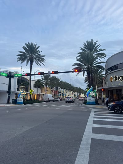 A street scene shows palm trees lining both sides of a road with cars parked along the edge. Traffic lights are red, and there's a pedestrian crosswalk. Buildings and businesses flank the road. Street signs indicate Harding Avenue and 96th Street. A bright blue and white sign labeled "SURFSIDE" with an illuminated sculpture is near the crosswalk. A building with the word "flag" is visible in the background.