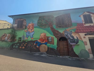 The mural, located on the building at Via Della Chiesa, Civita Castellana, Italy, depicts a scene from the fairytale "The Three Little Pigs." The mural features cartoonish pigs and a wolf painted on the side of a building. On the left, a pig in blue overalls holds a shovel, standing next to a small vegetable garden. In the center, a pig sits on a wooden barrel wearing a red shirt and blue overalls. To the right, a wolf is depicted near a wooden door. The mural is brightly colored and covers the entire facade of the building, extending to the roofline. The sky above is clear and blue. The building has windows with wooden shutters and a tiled roof. A road runs in front of the building.