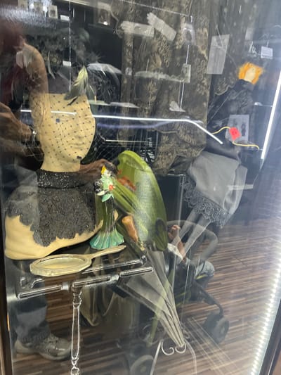 This image captures a display case in a shop, likely in Voskhod, Russia, showcasing vintage fashion accessories. The primary subject is a mannequin bust adorned with a veiled headdress and a heavily beaded black collar and bodice. To the right of the bust, a small figurine of a woman in a green dress holding flowers stands on a delicate stand. A polished silver hand mirror rests in front of the bust. In the background, various textiles with intricate patterns are visible, along with what appear to be framed photographs or cards. A bright green, somewhat abstract object is positioned behind the mannequin. The floor beneath the display is wooden and paneled. The overall impression is of a carefully curated collection of historical items.