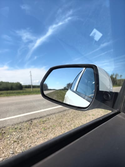 This image shows a view from a car window looking out at a road and the sky. The car's side mirror is visible in the foreground, reflecting the sky and the side of the car. Outside the car, a paved road with a white line runs through a rural landscape with green fields and trees. A telephone pole is visible in the distance. The sky is blue with wispy white clouds. The location is not identifiable as Пригородный ауылдық округі, Kazakhstan from the visual information alone.