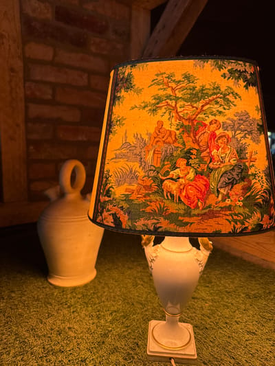 This media file captures a close-up, rotated view of a decorative table lamp with a lampshade featuring a pastoral scene. The lampshade is adorned with a tapestry-like print depicting figures in an idyllic outdoor setting, with trees, foliage, and architectural elements. The lamp base is a light-colored, urn-shaped ceramic, resting on a textured, carpet-like surface. The background reveals elements of an interior space, including exposed brickwork and wooden beams, suggesting a rustic or traditional decor. The lighting appears warm and ambient, possibly indicating evening or a dimly lit room. There is no visible text in the image. The overall impression is one of a cozy and artistically decorated corner of a room in Sankt Wendel, Germany.