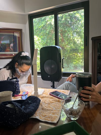 The image shows a scene inside a home in Río Oro, Costa Rica, with the coordinates 9.945 latitude and -84.201 longitude. A woman sits at a table, looking down at something on the table, while a second person's arm, likely the woman's, is extended toward the table. The scene is lit with natural light coming from a large window in the background, which shows lush green foliage. The woman wears a white shirt, dark pants, and a face mask. She has dark hair tied with a white ribbon. On the table are various objects, including a speaker, a cell phone, a cup, a bowl, a spoon, a towel, and a bag. The woman appears to be working on something on the table, possibly some sort of crafting project. The scene is peaceful and domestic.