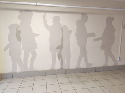 The image depicts a wall mural of light gray silhouettes of children on a beige wall. The silhouettes are arranged in a line, some facing each other, as if interacting. The figures vary in height and pose, with one figure raising an arm. The mural extends horizontally across the wall, with a tiled floor visible at the bottom. The wall is also adorned with a white pipe running along the top and an electrical outlet on the right side. This scene is located in Kurgan, Russia.