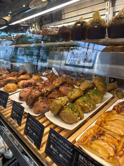 A bakery display case in Takarazuka, Japan, located at 34.809 latitude, 135.348 longitude. The case contains a variety of baked goods, including muffins, scones, pastries, and pies. The top shelf features four chocolate muffins with toppings and a price tag labeled "チョコレート ¥220." The second shelf holds an assortment of scones with various flavors, including chocolate, matcha, and fruit, displayed on a white tray with a price tag labeled "抹茶とチョコ ¥413." The bottom shelf features apple pie with a price tag labeled "国産りんごのアップルパイ ¥667." The bakery appears to be well-stocked and inviting.