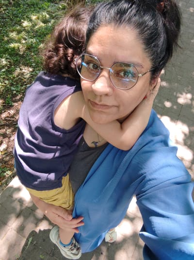 The image is a selfie taken from a high angle outdoors. A woman with dark hair pulled back, wearing large, polygonal glasses, is holding a young child who is facing away from the camera. The woman is wearing a dark tank top, a gray undershirt, and a bright blue scarf or shawl draped over her shoulder and arm. The child is wearing a dark purple t-shirt and yellow shorts with a pattern. The background shows a paved walkway with dappled sunlight filtering through trees, suggesting a park or outdoor public area. The ground is a mix of pavement and grass. There are no specific landmarks or indications that this location is in the city of Loris, Russia.