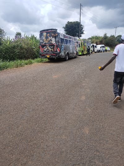 A row of Matatus lines a rural road, the first decorated with sports figures. A man in a white shirt and dark jeans walks by. The buses are parked on the road’s edge, with bushes and a field visible in the background. Power lines stretch across the cloudy sky. More vehicles are parked further down the road near people dressed in light-colored clothes. The road surface takes up a large portion of the frame.