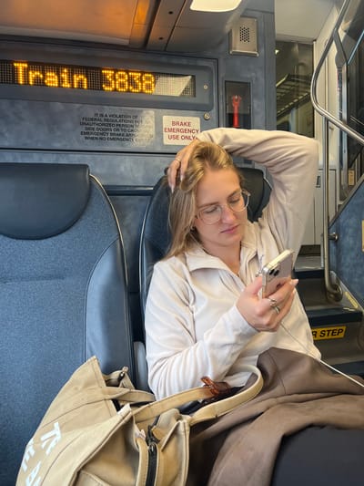 The image captures the interior of a commuter train, identified as "Train 3858" by an overhead LED display. Based on the signage and vehicle type common in the region, the location is a public transit train, consistent with NJ Transit services operating in or around Newark, United States.
A Caucasian female with blonde hair, wearing eyeglasses and a light beige quarter-zip fleece top, occupies a dark blue fabric seat. She is actively engaged with a light-colored smartphone held in her left hand, her gaze directed downwards at the screen. Her right arm is raised, with her fingers resting in her hair. Multiple rings are visible on her right hand. Adjacent to her on the seat or floor is a light tan canvas tote bag, upon which rests a small, dark brown, segmented wooden figurine. The train's interior features grey wall panels, a visible emergency brake pull handle, and informational signage regarding emergency brake usage and door operation, indicating "IT IS A VIOLATION OF FEDERAL REGULATIONS FOR UNAUTHORIZED PERSON TO OPEN SIDE DOORS OR TO CAUSE EMERGENCY BRAKE APPLICATION WHEN NO EMERGENCY EXISTS." A yellow-marked step with the partial inscription "NEXT STEP" is visible to the right. The passenger appears to be traveling alone and is focused solely on her device.
