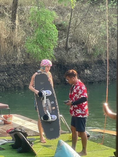 A woman wearing a pink polka dot helmet is being assisted onto a wakeboard on a dock. She is wearing wakeboarding bindings. A man wearing a red floral shirt is assisting her. The location is likely at or near the water in Tambon Nam Phrae, Thailand. The background includes trees, water, and a green artificial surface. A tow rope extends over the water.