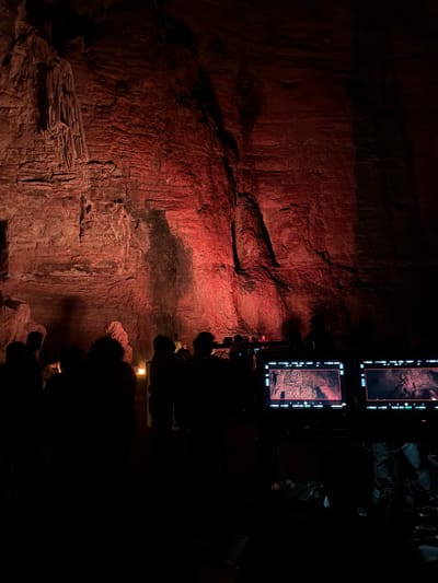 A group of people stand in silhouette in a dimly lit cave. The cave walls are rough and textured, illuminated by red lights that cast long shadows. In the foreground, two monitors display live camera feeds of the cave's interior, indicating a film or documentary production. The presence of cameras and monitors suggests that a recording or broadcast is taking place within the cave. The individuals appear to be observing the scene or participating in the production, their faces obscured by the low light conditions. The overall atmosphere is mysterious and dramatic, with the red lighting creating an intense visual effect. The location is identified as Gollagutta, India, a region known for its cave systems.
