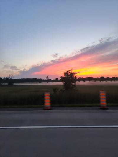 The image depicts a roadside view of a rural landscape at dusk. The sky is a gradient of soft blues transitioning to vibrant oranges and pinks, illuminated by the setting sun. Wispy clouds are painted with these sunset colors. In the midground, a field is covered in a low-lying mist that partially obscures a line of trees and silhouetted structures in the distance. The foreground features a section of asphalt road, distinguished by a white line. Two orange and white striped traffic cones are positioned on the shoulder of the road, indicating some form of traffic management or construction. The overall impression is one of tranquility and the natural beauty of twilight in a rural setting. The scene is likely captured from a moving vehicle, given the slight motion blur evident in the trees and cones. The location in Mirabel, Canada, suggests a setting characteristic of the Quebec countryside.