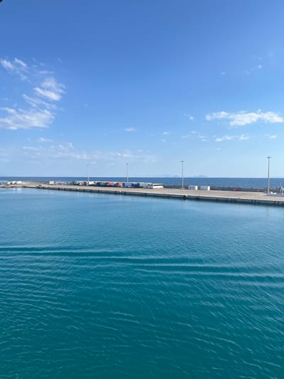 The image depicts a bright, clear day at a port. The sky is a vibrant blue with scattered white clouds. On the right side of the image, the deep blue water of the sea shows subtle ripples and patterns of light reflecting on its surface. A long, concrete pier stretches across the scene from left to right. Along the edge of the pier, numerous shipping containers are stacked and arranged. Several utility poles with lights are also visible along the pier. A few vehicles, including what appear to be trucks or vans, are present on the pier, suggesting active cargo operations. In the background, a distant shoreline with faint mountain outlines can be seen. The overall impression is one of a functional and busy port on a sunny day. The orientation of the image suggests it was taken from a slightly elevated vantage point, possibly from a boat or a higher structure overlooking the port. No people are clearly visible, and no text is discernible within the image. The scene appears to be set in the Municipal Unit of Kastro - Kyllini, Greece.