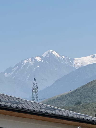 The image shows a view of a mountain range with snow-capped peaks under a clear blue sky. In the foreground, a tiled roof of a building is visible. A tall, lattice-like transmission tower stands in the middle ground, between the building and the mountains. The lower slopes of the mountains are covered in greenery. The image appears to be taken from a residential area in the vicinity of Belbulak, Kazakhstan, looking towards the mountains.