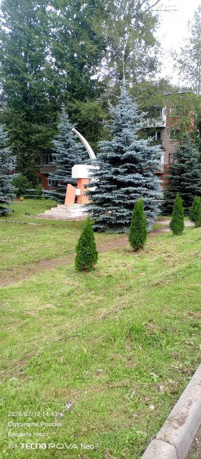 The image depicts an outdoor scene in Stroitel', Russia, featuring a monument situated on a grassy area with several trees. The monument is composed of a stepped base leading up to a sculpture that appears to be abstract, with orange and white geometric shapes, and a white curved element extending upwards. Surrounding the monument are various evergreen trees, including several prominent blue spruce trees, as well as smaller, conical green trees in the foreground. In the background, brick buildings with windows are visible, partially obscured by the trees, suggesting a residential or urban setting. The ground is covered in green grass, with some patches of dry leaves and debris. The lighting suggests it is daytime, possibly overcast. The timestamp "2026/07/12 14:43" and "Stroitel', Russia" are watermarked at the bottom left of the image, along with the text "Введите текст" (Enter text) and the TECNO POVA Neo 3 logo. There are no people visible in the image.