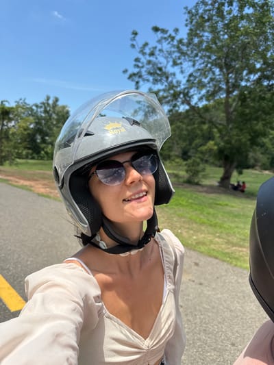A woman wearing a grey helmet with a clear visor and "Royal" logo is riding on a paved road in Ham Thuan Bac, Vietnam. She is wearing sunglasses and a light-colored top with a seashell necklace. The road has a yellow line marking on the left side. The background consists of lush green trees and grass under a clear blue sky. In the distance, a group of people can be seen sitting on the grass near a large tree. The image is a selfie, taken from the perspective of the rider.