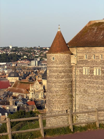 This is a high-angle, vertically oriented photograph of the town of Dieppe, France, viewed from a high vantage point. The foreground is dominated by the stone walls and red-tiled roofs of a large historical building, possibly a castle or cathedral, with a conical turret topped by a decorative finial. The stone masonry of the building is intricate, with numerous small, rectangular windows. Adjacent to the building, the scene transitions to a densely packed townscape with a variety of rooftops in shades of red, gray, and black, punctuated by church steeples and modern industrial silos in the distance. The town sprawls down a hillside towards a hazy horizon. To the right of the main building, a paved path runs alongside a grassy area enclosed by a low stone wall with metal fencing. The lighting suggests it is late afternoon or early evening, with the sun casting long shadows. The sky is clear and pale blue, indicating fair weather. No people are visible in the frame, and there is no discernible text.