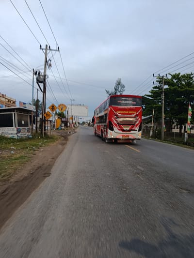 A red and white double-decker bus is traveling on a paved road in what appears to be a semi-urban or rural setting in Mesjid Tuha, Indonesia. The bus is the primary subject, viewed from the rear, and displays the text "HARAPAN INDAH" on its back. The road is gray asphalt with some patches of dirt and gravel on the shoulder. On the left side of the road, there are some buildings with signage, including one with the text "Always Low Prices." There are also several utility poles with electrical wires crisscrossing overhead. Yellow road signs, one depicting crossing pedestrians and another with a directional arrow, are visible near the roadside. Green trees and vegetation line the right side of the road, with some colorful flags planted in the ground. The sky is overcast and a dull gray, suggesting it might be dawn, dusk, or a cloudy day. The image is taken from a moving perspective, indicated by the motion blur on the road and surrounding elements. There are no people clearly visible in the immediate foreground or engaged in any specific actions.