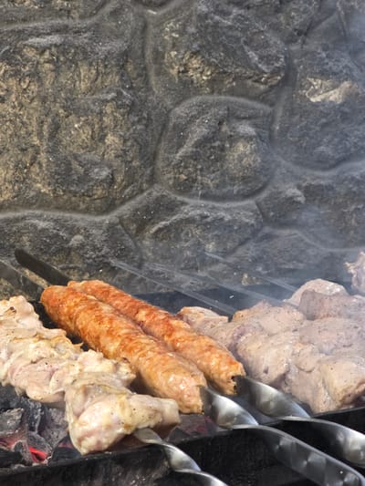 This media file captures a close-up, overhead view of food being grilled on skewers over hot coals. The foreground is dominated by the cooking meat, with several skewers visible. One skewer holds what appears to be sausages, which are golden brown and slightly glistening. Adjacent to the sausages are skewers with chunks of raw, seasoned meat, likely pork or chicken, with visible black pepper. A few pieces of the raw meat are already starting to brown on the edges from the heat. Wisps of smoke rise from the grill, obscuring parts of the scene and adding a dynamic element to the image.
The background features a rough, textured stone wall, likely a barbecue pit or outdoor kitchen structure. The stones are varied in color, ranging from dark gray to lighter shades of gray and tan, with a natural, uneven surface. The lighting suggests it is daytime, with natural light illuminating the food and the stone. There are no people or animals visible in the frame, focusing solely on the food preparation. The overall impression is one of a home-cooked meal or a casual outdoor gathering in Pidhorodne, Ukraine, centered around a traditional barbecue. The focus on the food and the grilling process evokes a sense of warmth and the anticipation of a delicious meal. No visible text is present in the image.