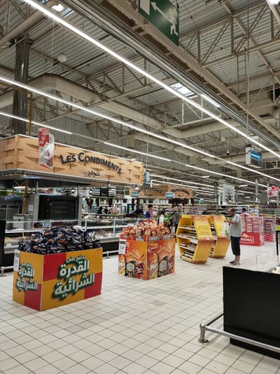 The image captures an indoor view of a brightly lit supermarket aisle, identified by signage as located in Cebalat, Tunisia. The ceiling is a complex network of metal beams and supports, with numerous fluorescent light fixtures illuminating the scene. Prominently displayed above a food counter is a large wooden sign with "LES CONDIMENTS" written in English and Arabic script. In the foreground, a large yellow and red display bin is filled with bags of Doritos chips, with Arabic text on the bin. Further into the aisle, various food products are on display, including snacks, jars of honey, and packaged goods. Several shoppers are visible, some browsing products and others walking through the aisle. A man in a grey t-shirt and dark shorts stands near a display of honey jars, holding a white plastic bag. The floor is tiled in a white grid pattern. The overall impression is of a busy, well-stocked retail environment.
