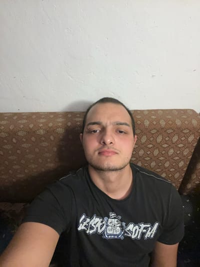 The image is a selfie taken indoors, likely in a residential setting. A young man with short dark hair, a light beard, and brown eyes is seated on a sofa with a patterned brown fabric. He is wearing a black t-shirt with white graphic text and imagery. The background consists of a white, textured wall. The lighting suggests an indoor environment, possibly during the evening or in a room with limited natural light. The man is looking directly at the camera with a neutral expression. The context of Dragoevo, Bulgaria, is not visually discernible from the image itself.