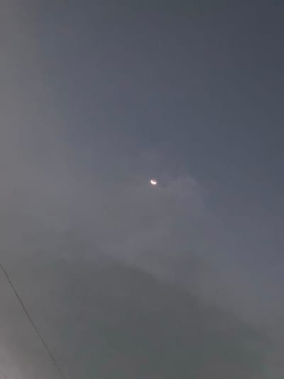 A crescent moon is visible in the overcast sky above Proyonangg, Indonesia. The sky is a muted gray, suggesting it is either dawn, dusk, or a cloudy day. The moon itself is a thin sliver, illuminated from the right. There are faint, wispy clouds partially obscuring the view of the moon. In the lower left corner of the image, a thin, dark line, possibly a power line, stretches diagonally across the frame. The overall impression is one of a subdued and calm atmosphere.