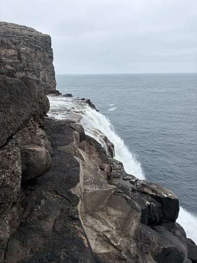The image shows a dramatic cliffside waterfall in Vatnsoyrar, Faroe Islands. The foreground is dominated by dark, rough-textured rocks, some of which are wet and glistening. Water cascades over the edge of the cliff, creating a white spray as it plunges into the sea below. The sea itself is a muted blue-grey, with a slight ripple pattern on its surface. The horizon is a soft, overcast grey sky. In the upper left corner of the image, the cliff face extends upwards, showing layers of rock. A few small, dark figures, likely people, are visible standing on the cliff edge in the distance. The overall impression is one of raw, natural beauty and a rugged coastal landscape.