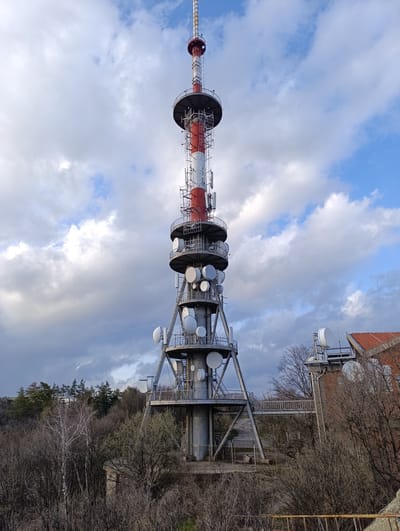 The image shows a tall telecommunications tower in Ichera, Bulgaria. The tower has a red and white striped mast extending vertically. The tower features several circular platforms with multiple dish antennas. The tower structure is supported by a metal framework. A building with a red tile roof is visible to the right of the tower base. The surrounding area is a mix of trees and vegetation. The sky is cloudy with patches of blue. The picture is taken from a slightly elevated position, looking up at the tower.
