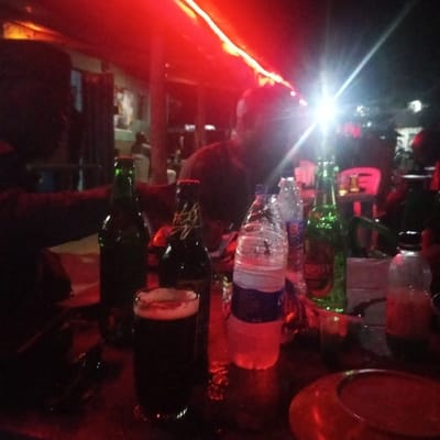 A low-light scene is depicted within an establishment, likely a bar or lounge, in Sauka, Nigeria. The environment is predominantly illuminated by red string or strip lighting overhead, casting a reddish hue across the area. Several individuals are present. In the foreground, two figures are seated at a table, largely silhouetted. One figure is positioned to the left, partially visible. Another figure, positioned right of center, is also a dark silhouette, with a very bright light source creating significant lens flare near their head.
The table in the foreground holds multiple beverage containers: a clear glass filled with a dark liquid (consistent with beer), at least two green glass beer bottles (one identifiable as "Trophy"), one dark glass beer bottle (partially identifiable as "Hero"), and several plastic water bottles, some containing water. A smaller clear bottle and a reddish-brown circular object, possibly an ashtray or dish, are also on the table. Additional blurred figures are visible in the dimly lit background.