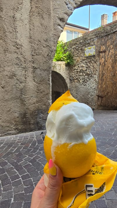 A close-up shot shows a hand holding a lemon filled with white ice cream, possibly a lemon sorbet or gelato, in an outdoor setting in Limone sul Garda, Italy. The lemon is cut in half, with the top half removed and placed atop the ice cream. The hand, with a yellow and pink gradient nail polish, grips the lemon. Behind the treat, a stone archway frames a view of a cobblestone street and buildings. A stone wall with a small sign adorned with sunflowers is visible on the right side of the archway. The scene suggests a warm, sunny day, likely in a historic European town, given the architecture and cobblestone streets.