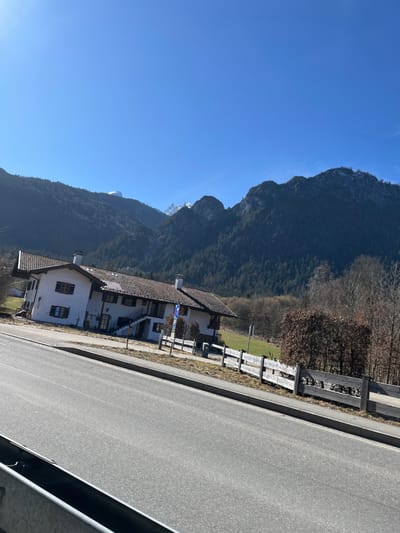 The image depicts a rural-mountainous landscape in Schönau am Königssee, Germany, under a clear blue sky. The scene is static, with no individuals present.
In the foreground, a two-lane asphalt road occupies the lower portion of the frame, angled diagonally upwards from left to right. A concrete curb separates the road from an unpaved shoulder or narrow strip of land. A metallic guardrail is partially visible in the extreme bottom-left corner.
In the midground, a two-story building with white facades and a dark reddish-brown tiled gabled roof is situated on a gentle slope to the right of the road. The building features multiple windows with dark-colored shutters. An external staircase with white railings leads to an upper-level entrance on the building's visible facade. Two chimneys protrude from the roofline. A blue and white circular road sign is mounted on a pole adjacent to the building. A light-colored wooden fence, composed of horizontal planks, runs parallel to the road, demarcating the property line. Behind the fence, a patch of green grass is visible, followed by a dense grouping of deciduous trees with bare branches and some shrubs with brown foliage, suggesting a non-summer season.
The background is dominated by a range of rugged mountains covered in dark green coniferous forests. Several peaks exhibit patches of snow, contrasting with the dark rock and tree-covered slopes. The sky above is clear and uniformly bright blue, devoid of clouds.