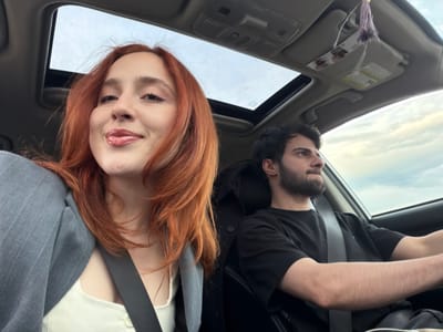 A car interior, viewed at an oblique angle through the open sunroof, shows a man and a woman. The man, on the left, has dark hair and a beard, and wears a black shirt. The woman, on the right, has long, red hair and is wearing a light-colored top and a gray blazer. Both are wearing seatbelts. The car’s interior includes the sun visor with an attached card and decorative tassels. The background visible through the sunroof suggests an outdoor environment with a cloudy sky. There is no indication that this image was taken in the city of Kvemo Nichbisi, Georgia.
