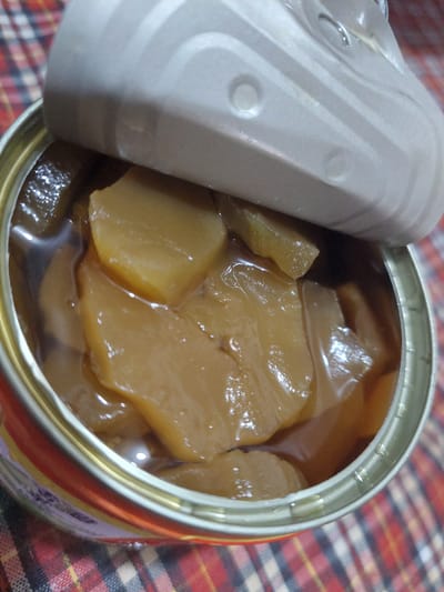 A close-up, high-angle shot shows the inside of an opened tin can filled with sliced, light brown fruit in syrup. The can's lid is partially open and to the upper right. The fruit slices are irregularly shaped and appear soft. The can is positioned on a red and white checkered surface. The scene is indoors, and no people are visible. The location in Jelutung, Indonesia is not discernible from the image.