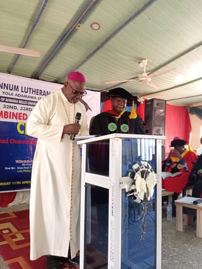 The image shows an indoor scene with a white podium decorated with a large black and white bow. The podium is positioned in front of a red and white banner that reads, “NNUM LUTHERAN, YOLA ADAMAWA ST, OF AHMADU BELLO UNIVER, 32ND, 33RD, COMBINED CH”. A man wearing a white robe and a purple hat is speaking into a microphone to the left of the podium. He is facing the podium. A man wearing a black cap and gown with a yellow tassel is seated behind the podium, facing the speaker. He is holding a book and looking down at the floor. There is another man seated to the right of him, partially visible. This person is also wearing a black cap and gown and has his head bowed. The ceiling of the room is tiled with wood panels. There is a ceiling fan in the background, as well as an open window. The image was taken at the coordinates 9.185 latitude and 12.521 longitude in the city of Rumde Waziri, Nigeria.