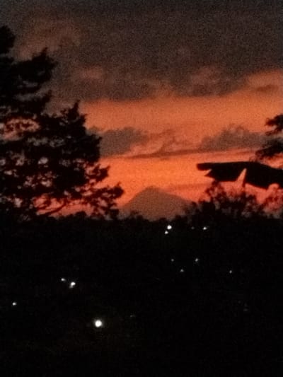 The image depicts a sunset scene with a mountain in the distance. The sky is a vibrant orange and red, with dark clouds obscuring some of the light. Silhouetted trees frame the left and right sides of the image, with a dark, shadowy foreground that suggests a landscape or hillside. In the distance, a conical mountain is visible against the colorful sky. The foreground is dotted with several small, bright lights, which could be distant houses or streetlights. The overall mood of the image is serene and somewhat dramatic, due to the intense colors of the sunset and the dark silhouettes. The time of day is clearly sunset. No people, objects, or text are clearly visible in the image. The location context is Menjing, Indonesia.