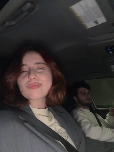 Two individuals are reclining in what appears to be the back of a vehicle. A woman with red hair and a slight smile is closest to the camera. A man with a beard is visible in the background. Both individuals seem to be wearing seatbelts. The interior of the vehicle is primarily gray. A white label is affixed to the vehicle’s interior on the left side of the image. There is no verifiable information in the image to confirm its location in the city of Grigalati, Georgia.