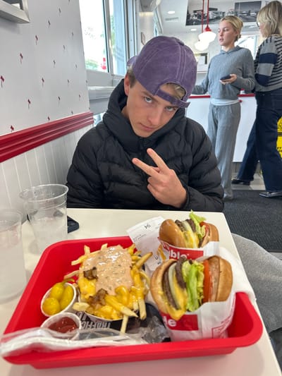 A young man wearing a purple baseball cap and a black puffer jacket poses with a peace sign. Beside him is a red tray containing two In-N-Out burgers wrapped in paper, a plate of animal-style fries topped with sauce, a small container of yellow peppers, ketchup, a spoon, and a napkin. Two empty plastic cups are on the table in the foreground. People are standing in the background, partially obscured. The table is white, and the wall is decorated with a red bird pattern.