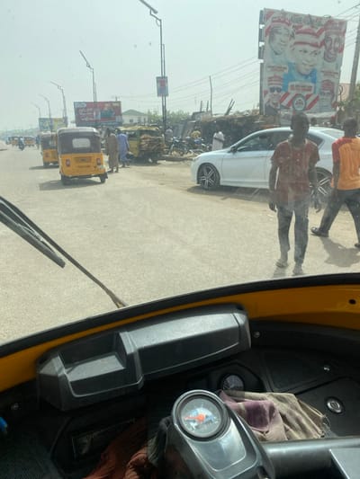 The image presents a daytime street scene in Gobirawa, Nigeria, captured from the driver's perspective inside a yellow motorized tricycle. The lower foreground shows the tricycle's dashboard, including a circular gauge on the steering column, a crumpled cloth, and the base of a windshield wiper.

Beyond the windshield, a paved, multi-lane road extends into the distance under a clear sky. On the left side of the road, several other yellow tricycles are present, some appearing to be in motion or parked. In the mid-ground on the right, a white sedan, resembling a Honda Accord with black and silver multi-spoke rims, is stationary or parked. Adjacent to it, a light-colored utility vehicle, heavily loaded with bundled wood or similar materials, is parked alongside several motorcycles.

Multiple individuals, all male, are visible on and near the roadway. Two men are prominent in the mid-right, walking away from the camera; one wears a faded red shirt, dark trousers, and sandals, while the other wears an orange shirt and dark trousers. Other men, some in traditional attire, are gathered near the loaded utility vehicle and further down the street.

The background on the right features a large billboard displaying portraits of five political figures and partial text including "ADAREANAU" and "PDP." Numerous streetlights equipped with solar panels line the road. Smaller billboards, utility poles, and distant buildings are also visible.
