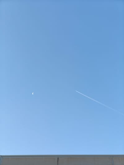 A clear blue sky dominates the upper portion of the image, with a crescent moon visible on the left side and a contrail from an airplane streaking across the right side. The lower portion of the image features a concrete barrier or wall, suggesting an outdoor or industrial setting. The image appears to be taken in Nàquera, Spain. There are no people or animals visible in the scene.