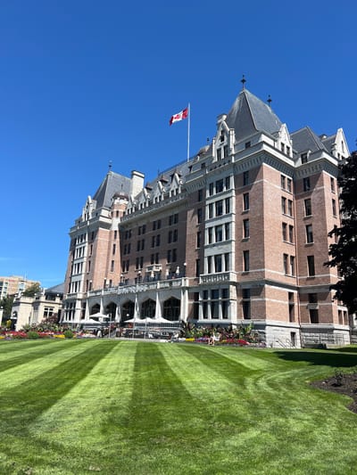 The image shows a large, historic hotel building with a Canadian flag flying on its roof, set against a clear blue sky. The hotel, identified by the sign "THE EMPRESS" above its entrance, is constructed of brick and stone with a distinctive architectural style featuring multiple gabled roofs and turrets. In front of the hotel, there is a meticulously manicured lawn with striped patterns created by mowing. A colorful flower bed with a variety of blooms runs along the base of the hotel. Several white umbrellas and outdoor seating areas are visible on a patio in front of the building, suggesting it is a public space, possibly a restaurant or cafe. A few people are seated at tables on the patio, and two individuals are visible in the flower bed. To the left of the hotel, another multi-story building with a sign indicating "HOTEL" is partially visible. The scene appears to be a sunny day in a well-maintained urban park or garden area. The Empress Hotel is located in Victoria, Canada, specifically on the Inner Harbour.