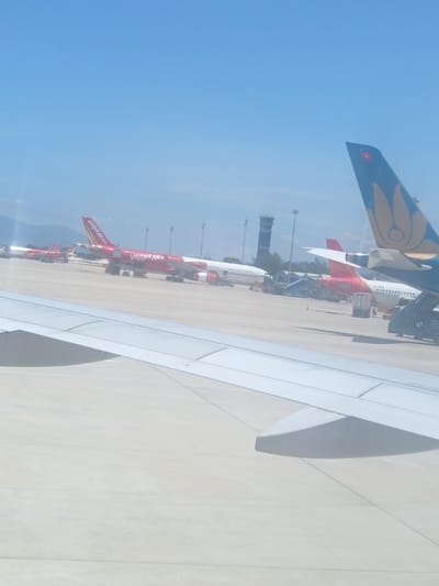 The image depicts an aerial view of Cam Ranh International Airport (CXR) in Cam Ranh, Vietnam, on a clear, sunny day. In the foreground, the wing of an airplane is visible, suggesting the photographer is on another aircraft. The tarmac of the airport is wide and appears to be made of concrete, with faint lines marking parking areas or taxiways.
Several airplanes are parked on the tarmac. Prominently featured are two red Vietjet Air airplanes, identifiable by their distinct livery with "Vietjet Air" written on the fuselage and "Vietjet.com" on the tail. One of these red planes is positioned closer to the foreground and has stairs attached to its side. Further back, more aircraft are visible, including what appears to be a white Vietnam Airlines plane with "PETROLIMEX AVIATION" written on it.
In the background, a tall, dark control tower stands out against the bright blue sky. Several lampposts are also scattered across the airport grounds. Mountains are faintly visible in the distance, behind the airport facilities. The overall scene conveys a busy, operational airport environment under a clear sky.
