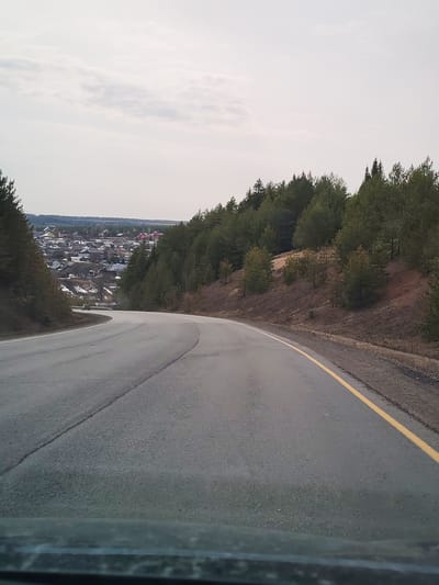 The image depicts a winding road in Doksha, Russia, with a view towards the town from an elevated position. The road is paved and has a yellow line marking the right edge. Evergreen trees line the right side of the road, while the town is visible to the left. The sky is overcast.